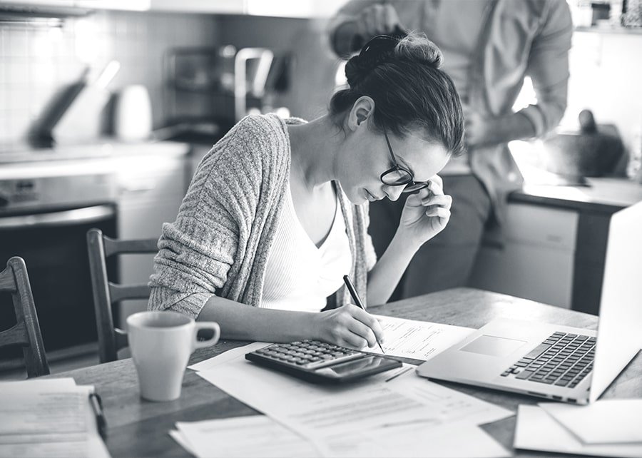 Woman at kitchen table with pen in hand looking down at paper with calculator and laptop nearby