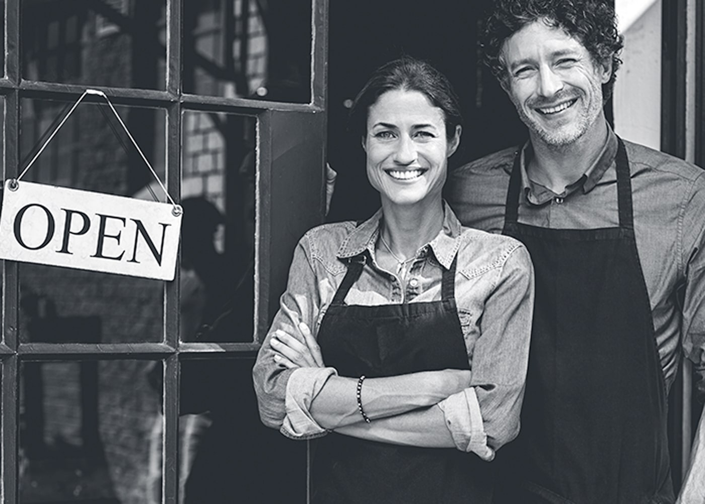 Smiling man and woman in aprons standing in front of business with open sign